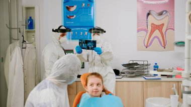 Koruyucu kıyafetli anne ve kızı klinikte pediatrik dişçi için bekliyorlar.