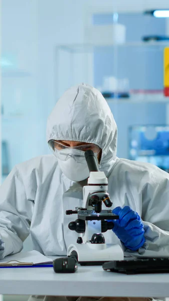 Biolog i ppe kostym arbetar med mikroskop och skriva information på Urklipp — Stockfoto