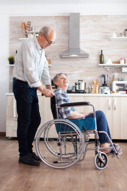 Mutlu koca, sakat yaşlı kadını mutfakta tekerlekli sandalyeye itiyor.