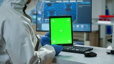 Koruyucu giysili kimyager elinde yeşil bir tablet tutuyor.