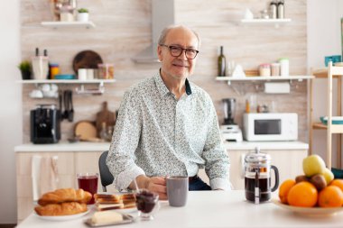 Mutfakta kıdemli bir adam elinde kahve fincanıyla kameraya bakıp gülümsüyor.