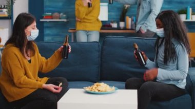 Kadınlar kanepede oturup koruyucu maske takıp abur cubur yemeyi tartışıyor.