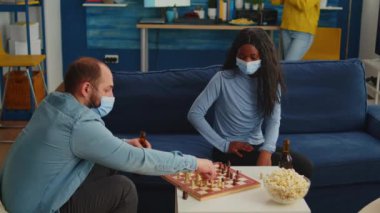Yüz maskesi takan Afrikalı bir kadın adamla satranç oynuyor.