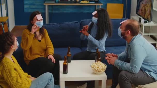 Olika vänner med ansiktsmask spelar spel med klibbiga anteckningar på forheads — Stockvideo