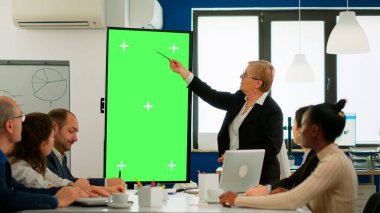 Takım lideri toplantı masasında durmuş mali istatistikleri analiz ediyor, yeşil ekrana bakıyor.