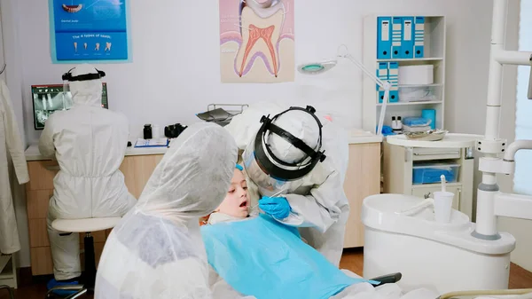 Pediatric dentist in protective shield checking teeth of little girl