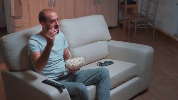 Caucásico macho sentado en un sofá con palomitas de maíz en las manos mientras mira la serie de películas — Vídeo de stock