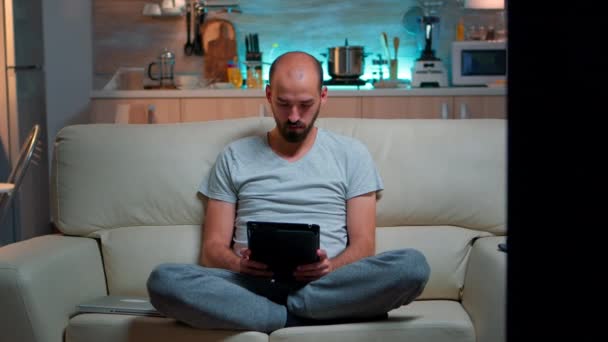Уставший человек сидит один на диване во время просмотра в Интернете с помощью планшетного компьютера с современной технологией беспроводной — стоковое видео