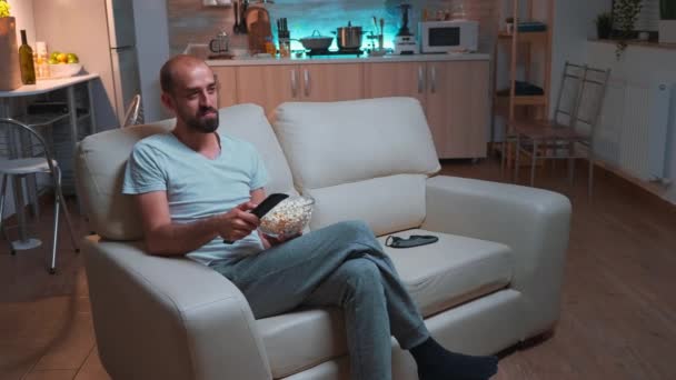 Homem com barba assistindo programa de tv usando controle remoto para mudar de canais na televisão — Vídeo de Stock