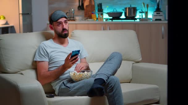 Скучный мужчина сидит на диване, держа чашу с попкорном — стоковое видео