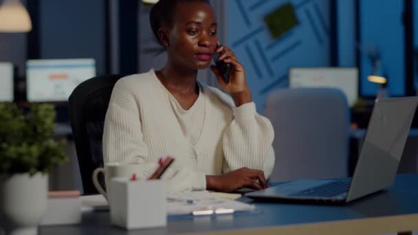 Empleado africano hablando por teléfono mientras trabaja en el portátil hasta tarde en la noche — Vídeos de Stock