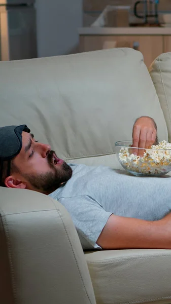 Hombre acostado en el sofá, comiendo popcron y viendo la televisión — Foto de Stock