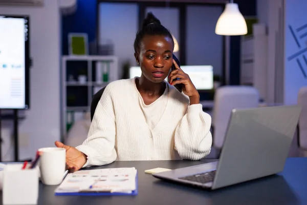 Африканський працівник розмовляє по телефону, працюючи в ноутбуці пізно ввечері. — стокове фото