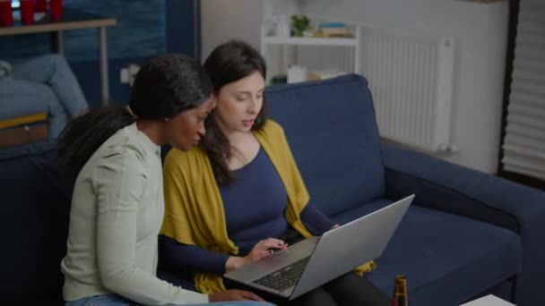 Twee multi-etnische vrouwen zitten op de bank te kijken comedy series op laptop — Stockvideo