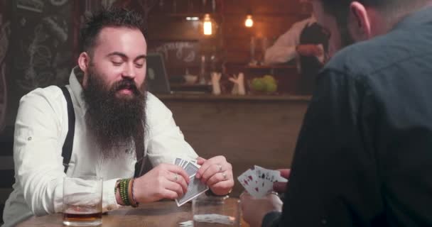 Positiver junger bärtiger Mann mit seinem Freund in einer Kneipe beim Kartenspiel — Stockvideo