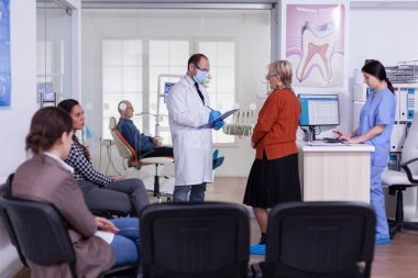 Maskeli bir ortodontist bekleme salonunda duran yaşlı bir kadınla konuşuyor.