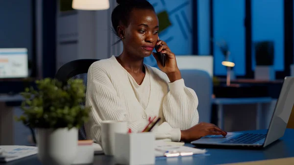 African employee speaking at phone while working at laptop late at night