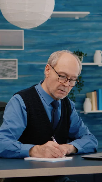 Multitasking retired man reading on laptop and writing on notebook — Stock Photo, Image