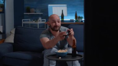 Heyecanlı odaklanmış bir adam kanepeye oturmuş elinde joystick ile video oyunları oynuyor.