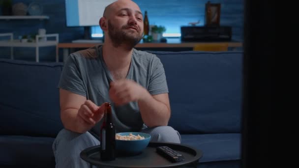 Mann sitzt allein auf gemütlicher Couch im Wohnzimmer, isst Popcorn, trinkt Bier — Stockvideo