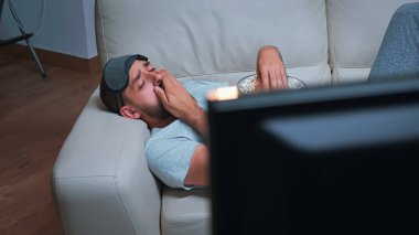 Beyaz erkek, göz maskeli, eğlence programı izlerken patlamış mısır yiyor.