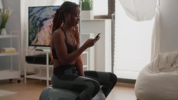 Λεπτή γυναίκα με σκούρο δέρμα κάθεται στο γυμναστήριο μπάλα γράφοντας μήνυμα στο τηλέφωνο — Αρχείο Βίντεο