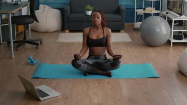 Esnek, ince, siyah kadın oturma odasında yoga haritasında lotus pozisyonunda oturuyor.