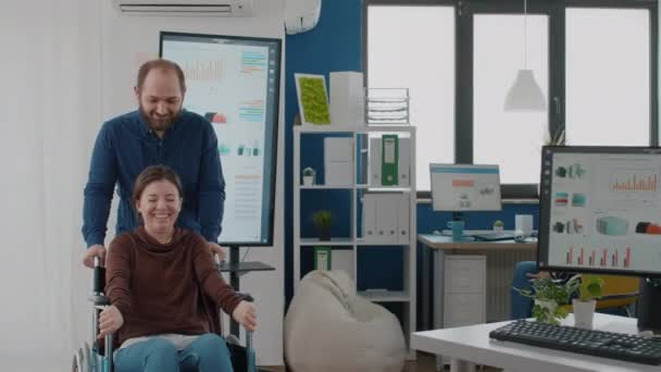 Joven alegre empujando silla de ruedas con su compañero de trabajo paralizado discapacitado — Vídeos de Stock