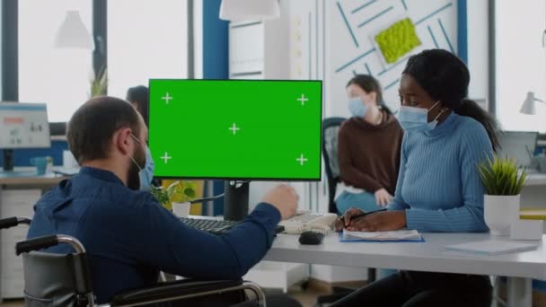 Manager mit Gesichtsmaske tippt auf PC mit grünem Bildschirm diskutiert mit afrikanischem Mitarbeiter — Stockvideo