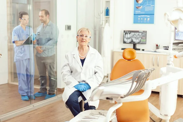 Стоматолог пожилая женщина смотрит в камеру в ожидании больного мужчины — стоковое фото