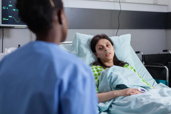 Nemocná hospitalizovaná žena ležící v posteli, zatímco afro-americká zdravotní sestra kontrolující příznaky onemocnění — Stock fotografie