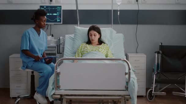 Взрослая больная женщина отдыхает в постели, в то время как черная медсестра анализирует болезнь — стоковое видео