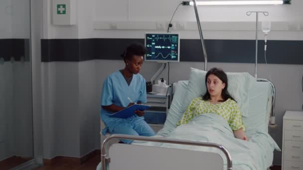 Sick woman resting in bed while afro-american nurse monitoring disease — Stock Video