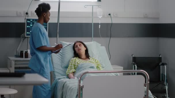 Afro americká asisstant kontrola nemocné ženy, zatímco sedí v posteli v nemocnici — Stock video