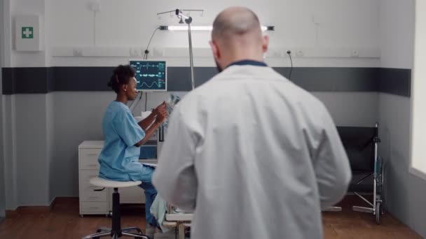 Afro-amerikan hemşire röntgen kemiklerini incelerken hasta kadının yatakta yatışı ön planda. — Stok video