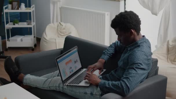 Afro-Amerikaanse man werkt vanuit huis, werkt op afstand op laptop computer met online grafieken en statistieken — Stockvideo
