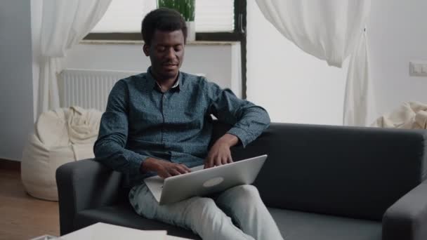 Relajarse en el sofá, chico afroamericano escribiendo y trabajando con un ordenador portátil moderno — Vídeo de stock