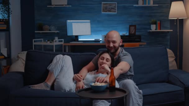 Piękna szczęśliwa para ogląda telewizję na kanapie relaksując się w nocy śmiejąc — Wideo stockowe