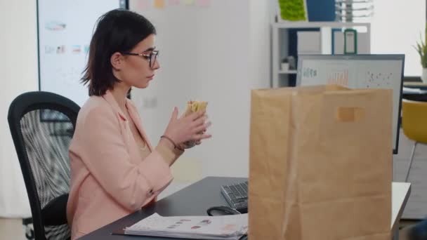 Empresaria comiendo bocado de sabroso sándwich bebiendo café delante del monitor — Vídeos de Stock