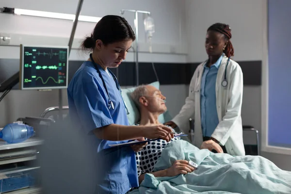 Assistente medico che prende appunti sugli appunti mentre il medico africano rassicura il paziente anziano — Foto Stock