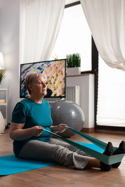 Focused pensioner wearing sportswear sitting on yoga mat stretching legs muscle — Stock Photo, Image