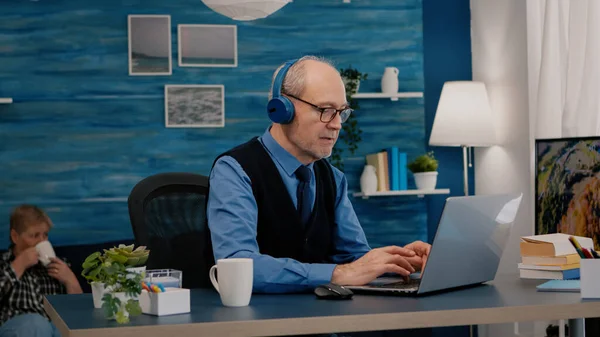 Vecchio imprenditore concentrato con cuffie ascoltare musica digitando sul computer portatile — Foto Stock