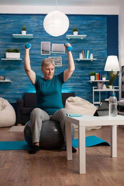 Senior woman in retirement raising hands doing arms exercise using dumbbells sitting fitball — Stock Photo, Image