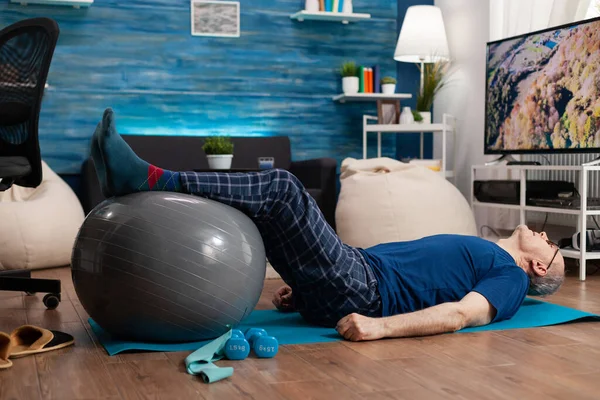 Jubilación senior hombre haciendo bienestar calentando piernas hasta usando swiss ball sentado en yoga mat —  Fotos de Stock