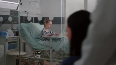Hastanede oksijen tüpü takan hasta bir kız yatağında oturmuş dizüstü bilgisayarında video oyunları oynuyor.