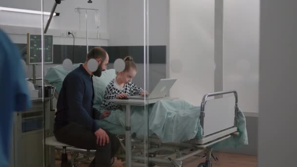 Padre sentado con la hija enferma en la sala de hospital mientras juega videojuegos de terapia en línea con el ordenador portátil — Vídeos de Stock