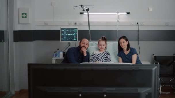 Nemocný hospitalizovaný dětský pacient odpočívající v posteli s rodinou sledující kreslený film v televizi — Stock video