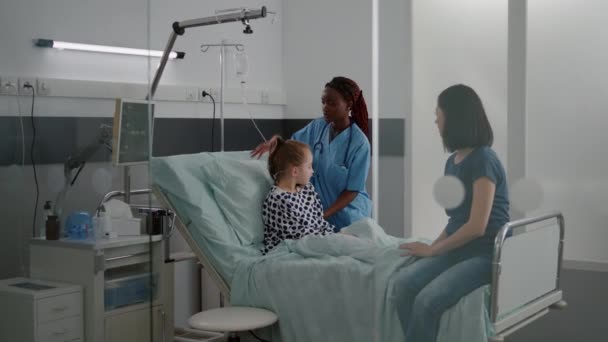 Мать обсуждает с больной маленькой дочерью в то время как африканская американская медсестра организовать кровать — стоковое видео