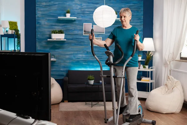 Invalid senior woman doing aerobics on cycling bike machine in living room for well being — Stock Photo, Image
