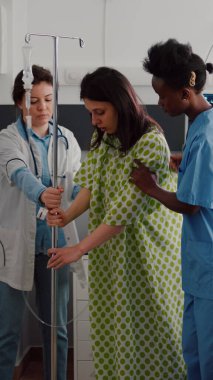 Tıbbi ekip hastanın yataktan kalkmasına yardım ediyor. Damardan damar içi sıvı akışı torbası tutuyorlar.